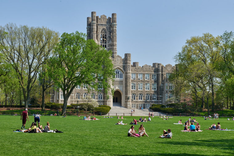 Old stone building with students outside lounging on a green lawn.