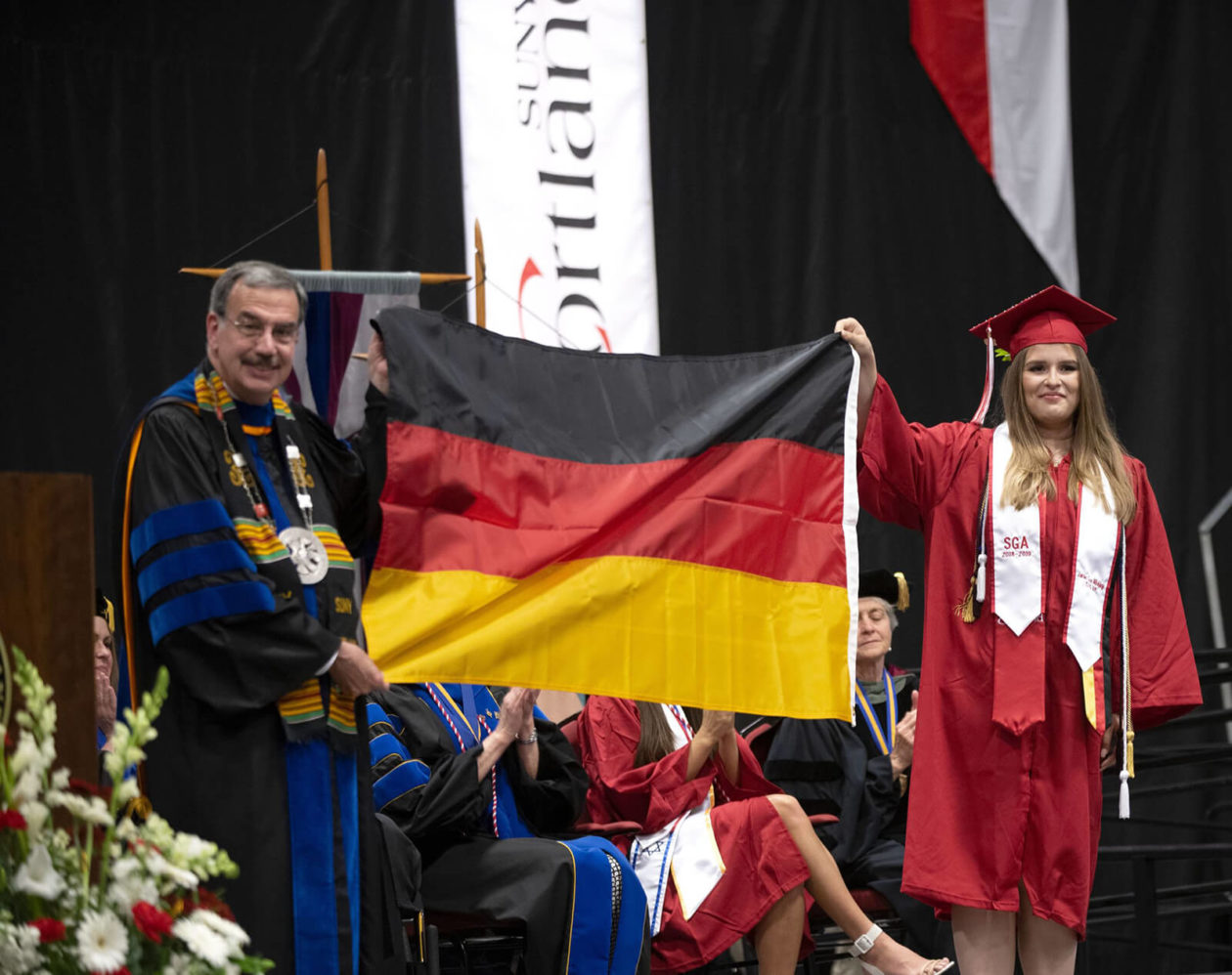Student in a red graduation cap and gown holding a flag.
